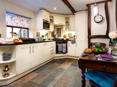 A kitchen with white cabinets a tiled floor a wooden dining table with place settings and food and a window with patterned blind at Tipsy Gin Cottage in Crosthwaite