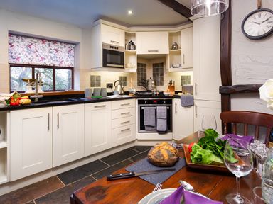 A kitchen with white cabinets a black countertop a microwave an oven and a dining table with bread and salad at Tipsy Gin Cottage in Crosthwaite