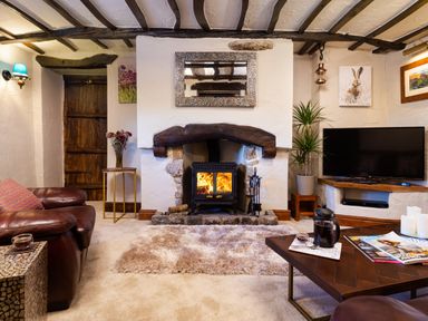 A living room with a wood burning stove in a stone fireplace a brown leather sofa a television and wooden beams on the ceiling at Tipsy Gin Cottage in Crosthwaite