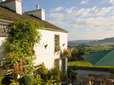 An outdoor seating area with wooden tables and chairs next to a white building covered with plants and greenery with a green umbrella and countryside view at Tipsy Gin Cottage in Crosthwaite