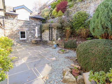 A stone patio with outdoor table and chairs surrounded by stone walls and shrubs at Courtyard Cottage in Ambleside