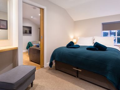 A bedroom with a bed covered with a blue blanket and rolled towels next to a window and a sliding door leading to a living area with a TV at Barn End Ambleside