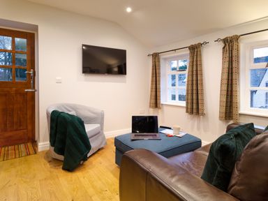 A sitting room with a brown leather sofa green cushions blue ottoman with a laptop and cup a grey armchair with a green blanket and a wall mounted television at Barn End in Ambleside