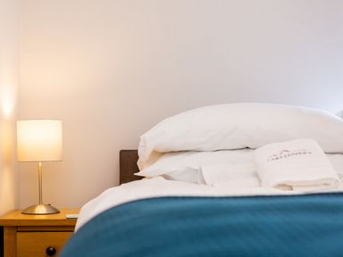 A bedroom with a bed with white pillows and towels and a blue blanket next to a wooden nightstand with a lamp at Barn End in Ambleside
