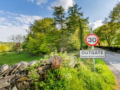 A country road with a 30 mph speed limit sign and an outgate sign next to a stone wall and trees at Barn End in Ambleside