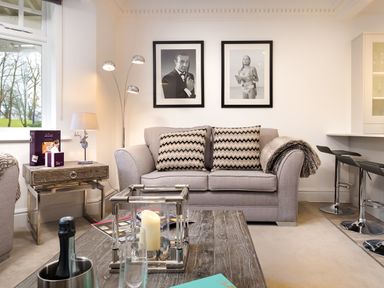 A living room with a gray sofa and two patterned cushions two black and white pictures on the wall a wooden coffee table with candles a side table with a lamp and a window in Skyfall in Bowness