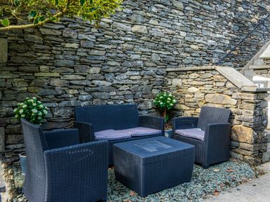 An outdoor seating area with black wicker furniture and cushions against a stone wall at Skyfall in Bowness