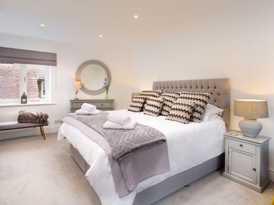 A bedroom with a large bed with patterned pillows bedside tables with lamps a bench near a window and a dresser with a round mirror at Skyfall in Bowness