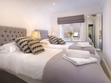 A bedroom with two single beds lamps a window with a vase of flowers and a wall mirror at Skyfall in Bowness
