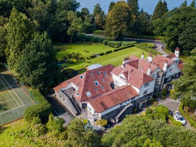 An aerial view of a large house with red roofs surrounded by trees and greenery with a tennis court and a lake in the background at Skyfall in Bowness