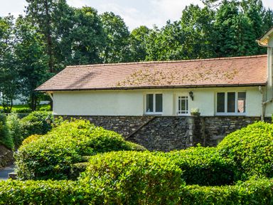 A stone and white plaster building with a tiled roof surrounded by green bushes and trees at Skyfall in Bowness