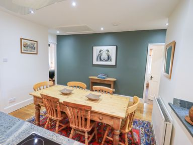 A dining room with a wooden table and six chairs on a patterned rug and artwork on the wall at Rowan Cottage in Grasmere