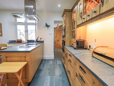 A kitchen with wooden cabinets a countertop with a coffee machine and bread box and an island with a stove and extractor fan at Rowan Cottage in Grasmere