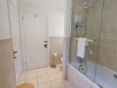 A bathroom with a bathtub shower combination along the right wall a toilet with a freestanding toilet paper holder and two closed white doors Rowan Cottage Grasmere