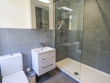 A bathroom with a toilet, a sink with drawers, a trash bin, and a glass-enclosed shower at Rowan Cottage in Grasmere