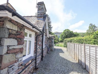 Exterior side view of a stone cottage with a gravel pathway and wooden fence at Rowan Cottage in Grasmere