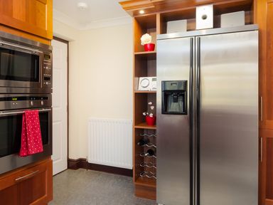 A kitchen corner with a stainless steel double door refrigerator a built-in microwave and oven a red towel on the oven handle wooden shelves with a wine rack and decorative items