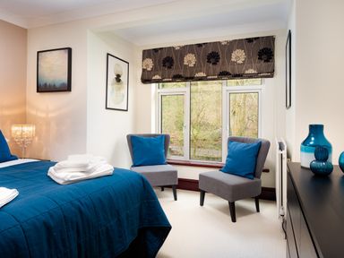 A bedroom with a blue bedspread two gray chairs with blue cushions and a window with floral blinds at Beech View in Ambleside