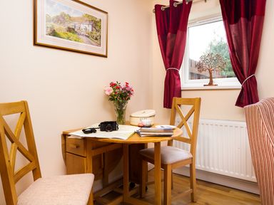 A small dining area with a wooden table and chairs a vase with flowers on the table and red curtains by the window at Artists Hideaway in Keswick