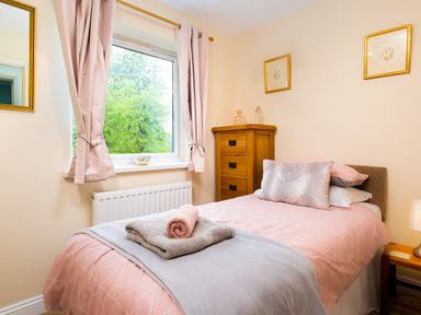 A bedroom with a single bed with pink bedding a wooden chest of drawers and a window with curtains at Artists Hideaway in Keswick