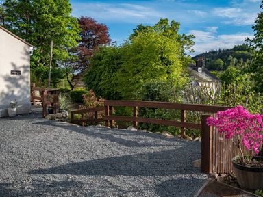 A gravel driveway with wooden fencing and potted pink flowers surrounded by trees at Artists Hideaway in Keswick