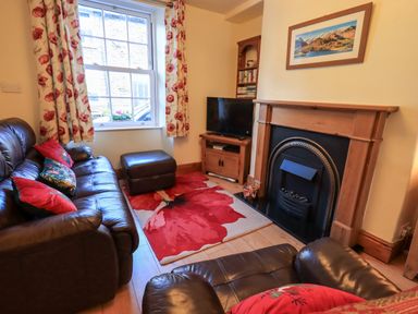 A sitting room with leather sofas a fireplace a television floral curtains and a red floral rug at Tizzie Whizie Cottage in Windermere