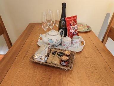 A wooden dining table with a tea set champagne bottle four glasses a packet of chocolates and a basket with coffee and jam jars at Tizzie Whizie Cottage in Windermere