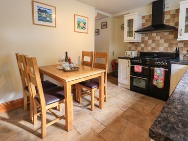 A kitchen with a wooden dining table and four chairs and a black stove with oven mitts at Tizzie Whizie Cottage in Windermere