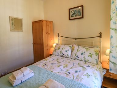 A bedroom with a double bed with floral bedding a wooden wardrobe bedside tables with lamps and folded towels on the bed at Tizzie Whizie Cottage in Windermere