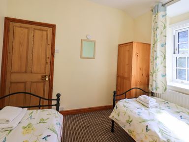 A bedroom with two single beds a wooden door a wooden wardrobe and a window with floral curtains at Tizzie Whizie Cottage in Windermere