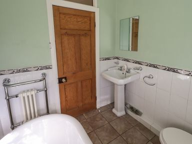 A bathroom with a wooden door a white pedestal sink a mirror and part of a bathtub at Tizzie Whizie Cottage in Windermere