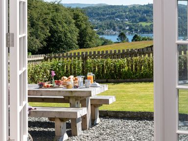 A picnic table set with breakfast items on a gravel patio with a lawn and fenced garden overlooking a lake and hills at Hawkrigg Farm in Hawkshead