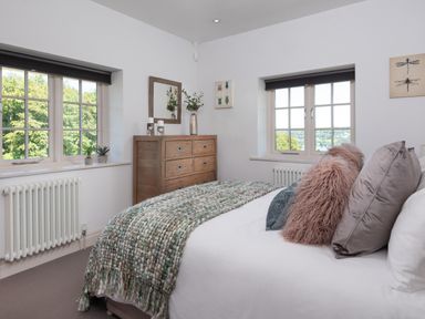 A bedroom with a bed with pillows and a blanket a wooden dresser with a mirror and plants and two windows overlooking greenery at Hawkrigg Farm in Hawkshead