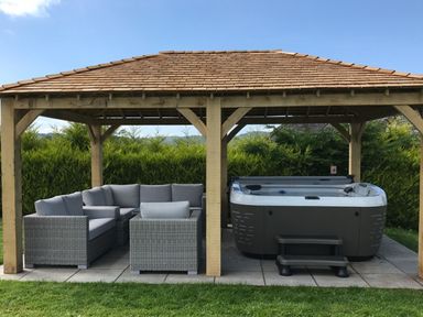 An outdoor wooden gazebo with a grey cushioned seating area and a hot tub on a tiled floor surrounded by green bushes at Hawkrigg Farm in Hawkshead