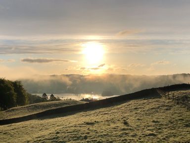 A grassy hillside with a stone wall and morning sun over a misty lake at Hawkrigg Farm in Hawkshead
