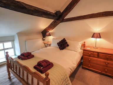 A bedroom with a wooden bed with white bedding and burgundy towels next to wooden nightstands with lamps under wooden ceiling beams at Blease Garth in Threlkeld