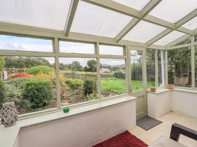 A sunroom with large windows overlooking a garden with bushes and trees at Blease Garth in Threlkeld