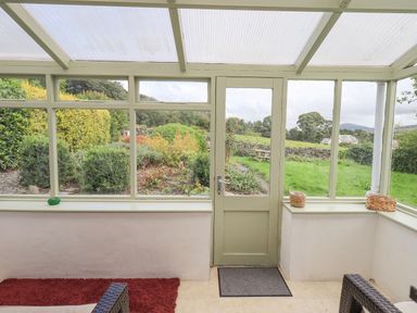 A sunroom with glass windows and door overlooking a garden with bushes and a picnic table at Blease Garth in Threlkeld