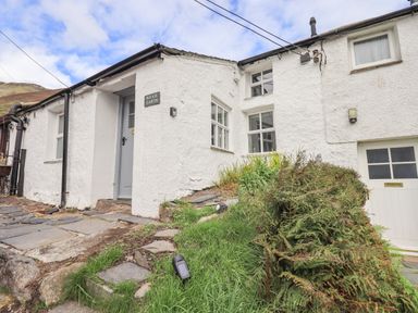 The exterior of a white painted cottage with a gray door and stone pathway surrounded by grass and plants at Blease Garth in Threlkeld