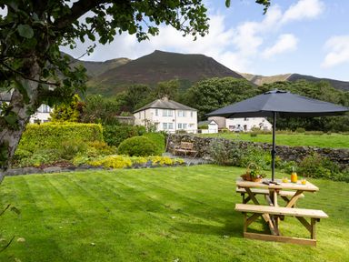 A garden with a wooden picnic table and umbrella and houses with hills in the background at Blease Garth in Threlkeld