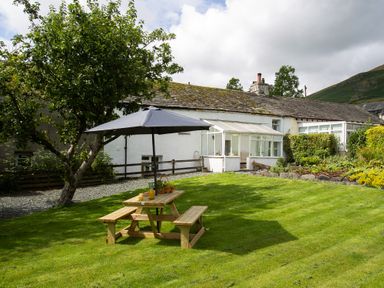 A garden with a wooden picnic table and umbrella in front of a white house with a glass porch at Blease Garth in Threlkeld