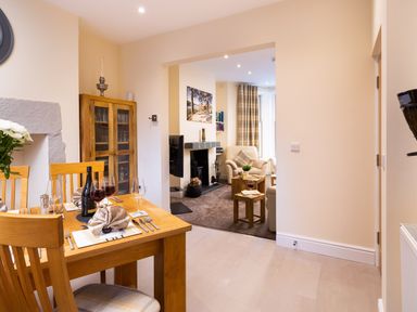 A dining area with a wooden table set for four with wine glasses and a bottle and an adjoining living room with armchairs and a fireplace at Dovedale Keswick