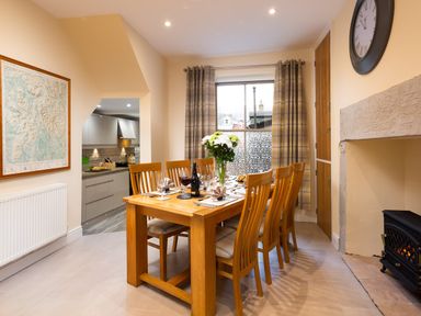 A dining room with a wooden table and chairs a vase with flowers on the table a wall clock a radiator and a view into a kitchen at Dovedale in Keswick
