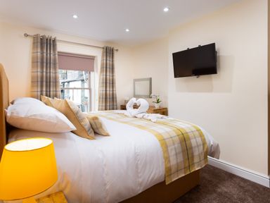 A bedroom with a bed covered in a plaid blanket pillows and two towel swans a window with plaid curtains a wooden chest of drawers and a wall-mounted television at Dovedale in Keswick