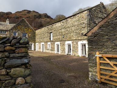 A stone walled building with multiple white framed doors and windows and a wooden gate in the foreground at Kentmere Hall Bank Barn in Kentmere