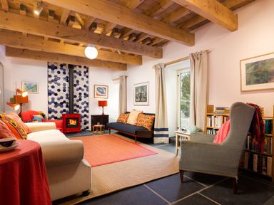 A living room with wooden ceiling beams red stove blue couch beige armchair and bookshelves at Kentmere Hall Bank Barn in Kentmere