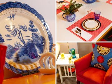 A blue and white decorative plate with a scenic design a dining table set with a plate glass and cutlery on a red placemat and a blue and white vase with green foliage and a red chair with a patterned pillow and a small white side table with glasses and a blue pitcher