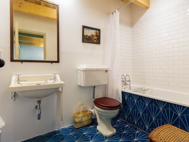 A bathroom with a wall-mounted sink under a mirror a toilet with a brown seat a bathtub with white tiles and blue geometric patterned lower panels and a wicker basket on the floor at Kentmere Hall Bank Barn Kentmere