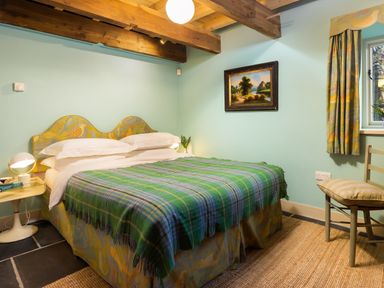 A bedroom with a double bed covered by a green plaid blanket a nightstand with a lamp a chair with a cushion a framed painting on the wall and a window at Kentmere Hall Bank Barn in Kentmere