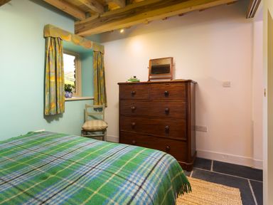 A bedroom with a bed covered with a green plaid blanket a wooden chair with a cushion a window with patterned curtains and a wooden dresser with a mirror on top at Kentmere Hall Bank Barn in Kentmere
